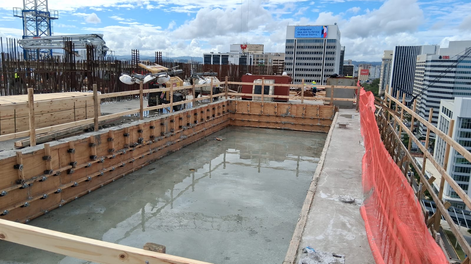 Piscina comercial en construcción en una azotea, con encofrado y hormigón, frente al skyline de San Juan